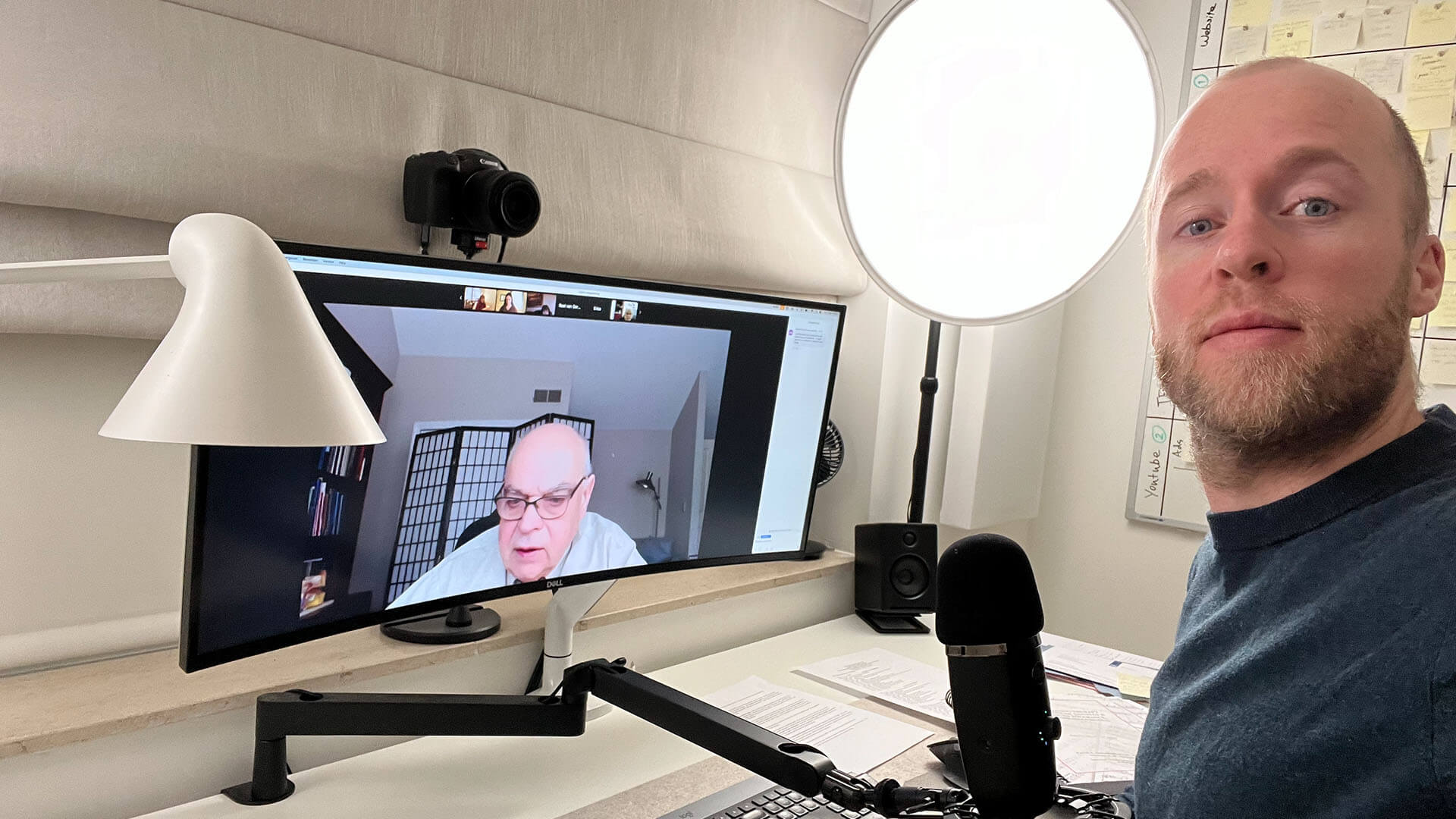 Tinnitus expert Roel van Gorkum sitting at his desk during a video call with tinnitus expert Dr. Jastreboff on a large screen; professional podcasting setup with mic, ring light, and camera.