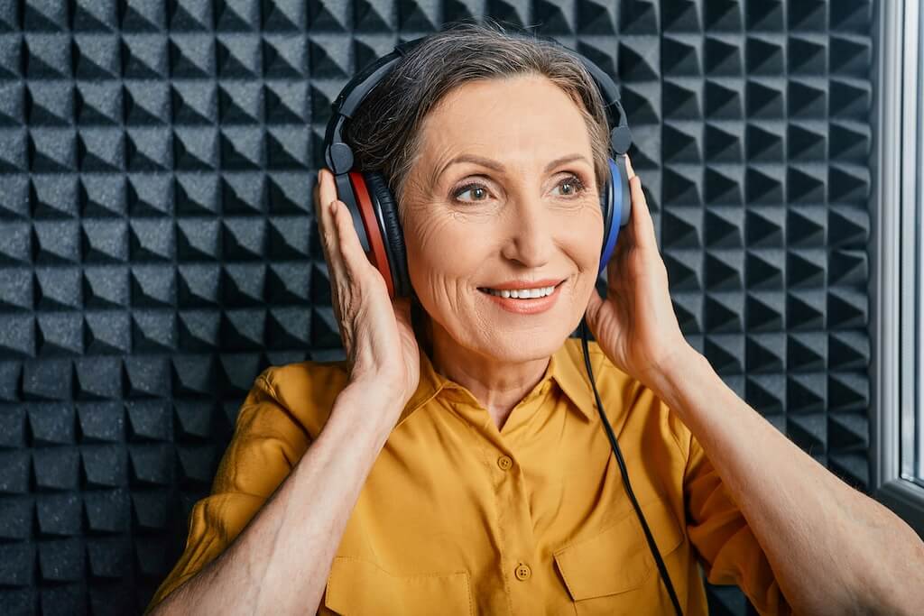 A smiling older woman wearing headphones in a soundproof studio.