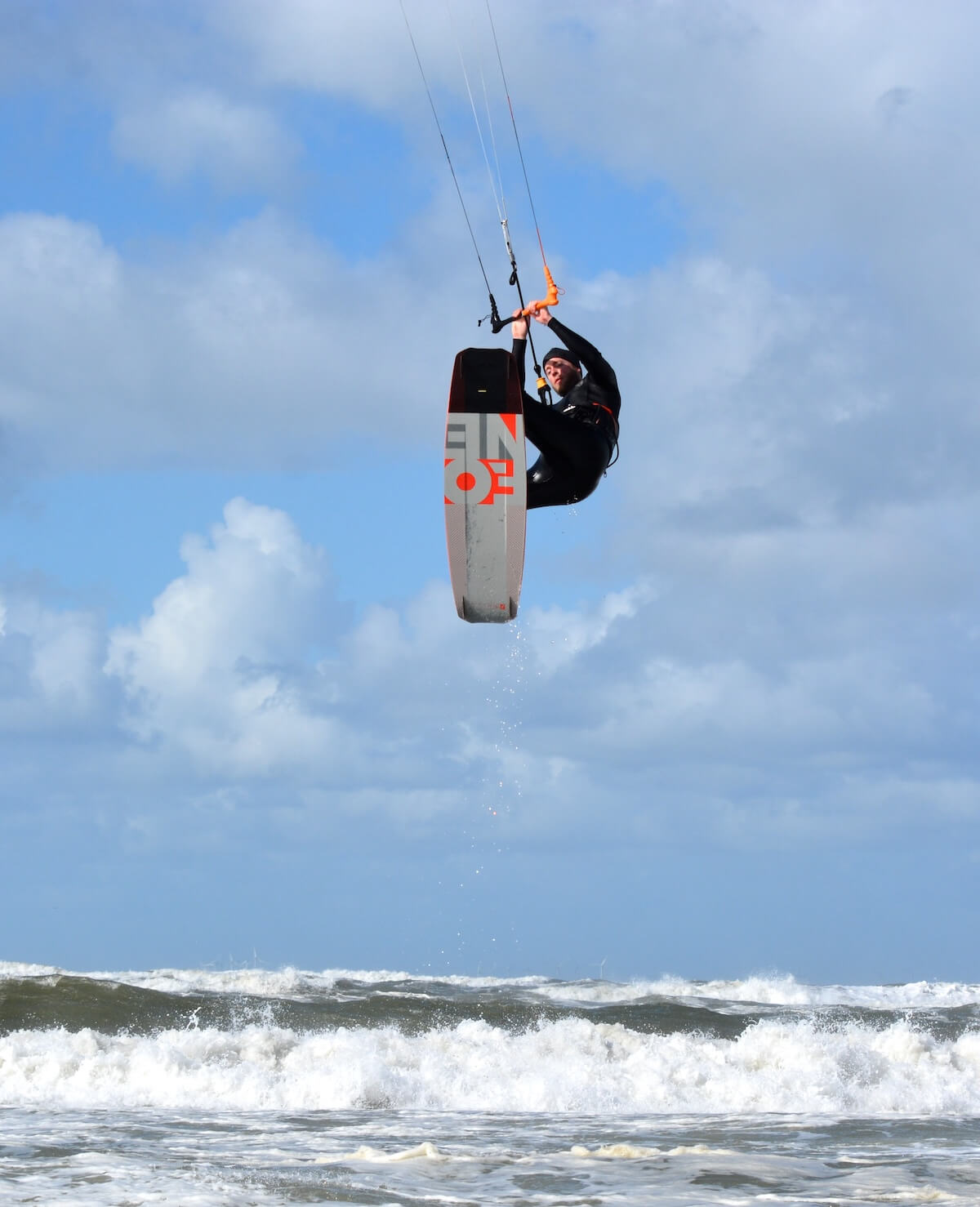 Roel van Gorkum springt tijdens het kitesurfen boven de oceaangolven uit op een zonnige dag met verspreide wolken.