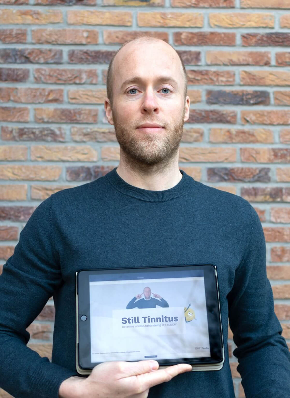 Roel van Gorkum, Still Tinnitus founder, stands in front of a brick wall, holding a tablet displaying the text "Still Tinnitus."