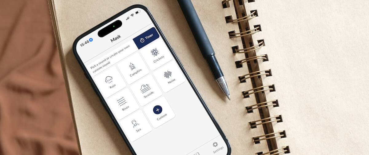 Een smartphone waarop de Still Tinnitus-app wordt weergegeven met geluidsopties (regen, kampvuur, krekels, enz.), geplaatst op een notitieboek met daarnaast een pen.