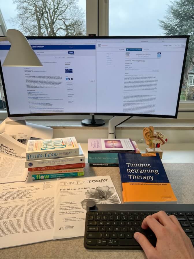 Een werkplek met een computermonitor waarop medische artikelen worden weergegeven, een toetsenbord en verschillende boeken over mentale gezondheid en tinnitus­therapie. Er liggen ook verspreide papieren en er is een klein anatomisch model aanwezig.
